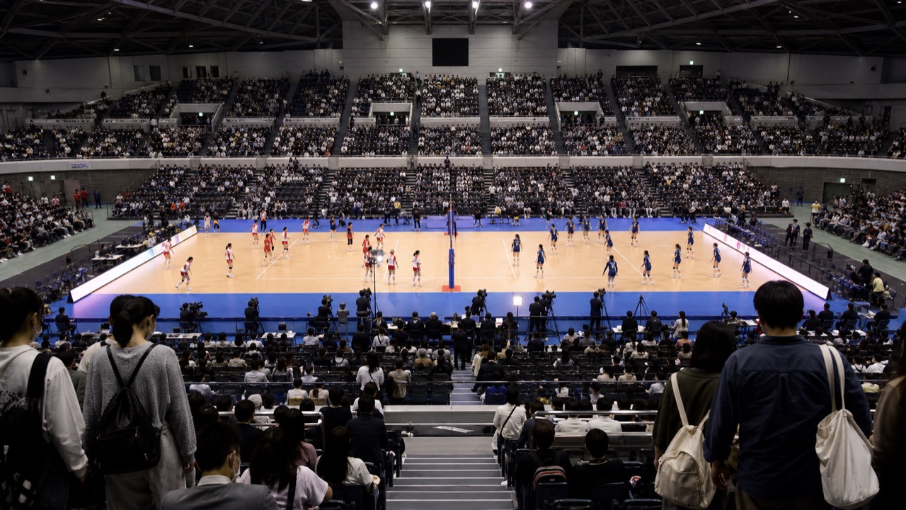 満員の屋内アリーナで行われるバレーボールの試合を客席後方から見た編集イメージ